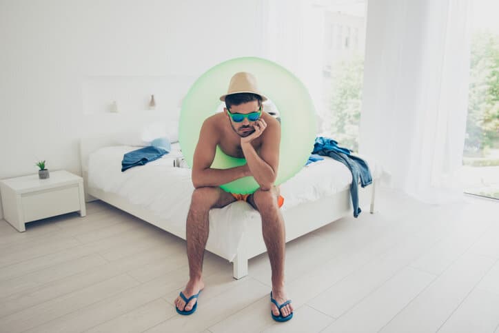 Attractive handsome young bearded sad brunet man wearing swim wear, life buoy, sunglasses, sitting indoors on bed. Summer vacation at sea resort
