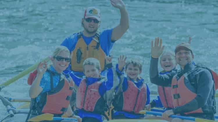 Group of adults and kids smiling and waving to the camera from a raft on the Yellowstone River.